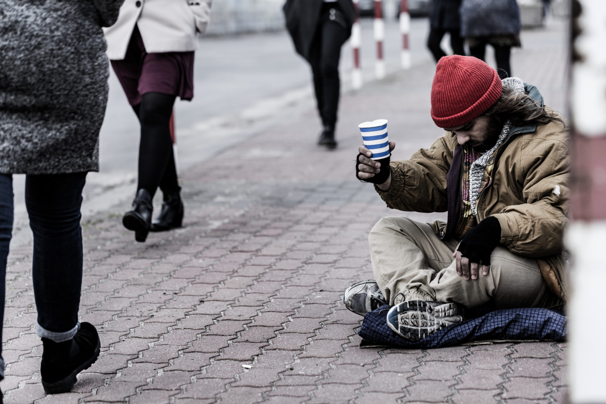Hopeless beggar asking for food while sitting on the sidewalk between pedestrians